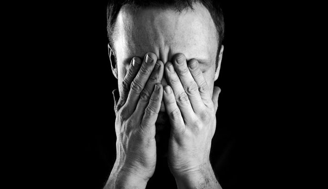 Monochrome portrait of young stressed Caucasian man covers his face with hands isolated on black background