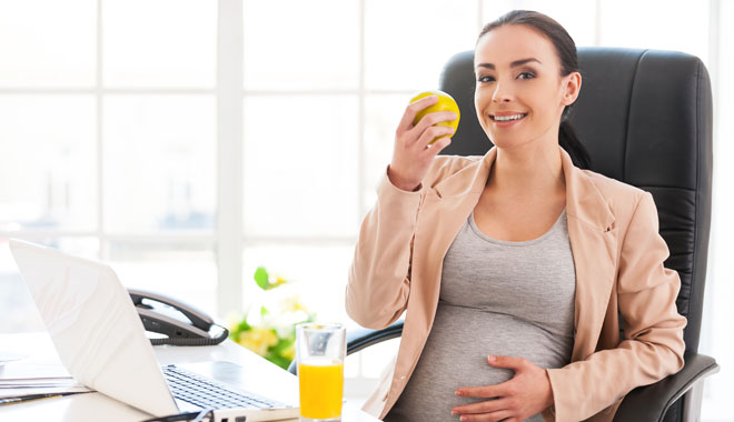 Pregnant woman in office. Beautiful pregnant businesswoman eating apple and smiling at camera while sitting at her working place