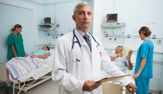 Doctor with clipboard in busy hospital room with two nurses talking to patients in background
